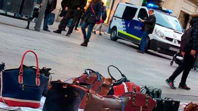 Bolsos a la venta en un 'top manta' en Barcelona junto a una furgoneta de la Guardia Urbana.