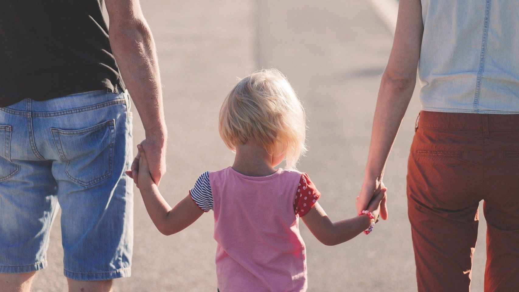 Una niña camina cogida de las manos de sus padres / CG