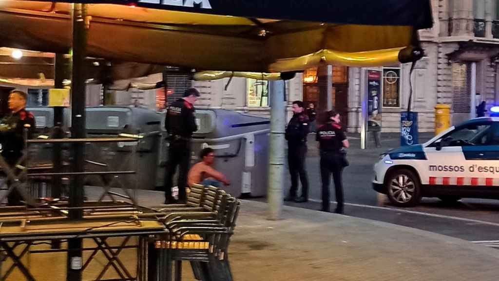 Una de las dos personas involucradas en la pelea (centro) sentada en una acera de la plaza de Cataluña acompañada de los Mossos d'Esquadra / HUGO SÁNCHEZ