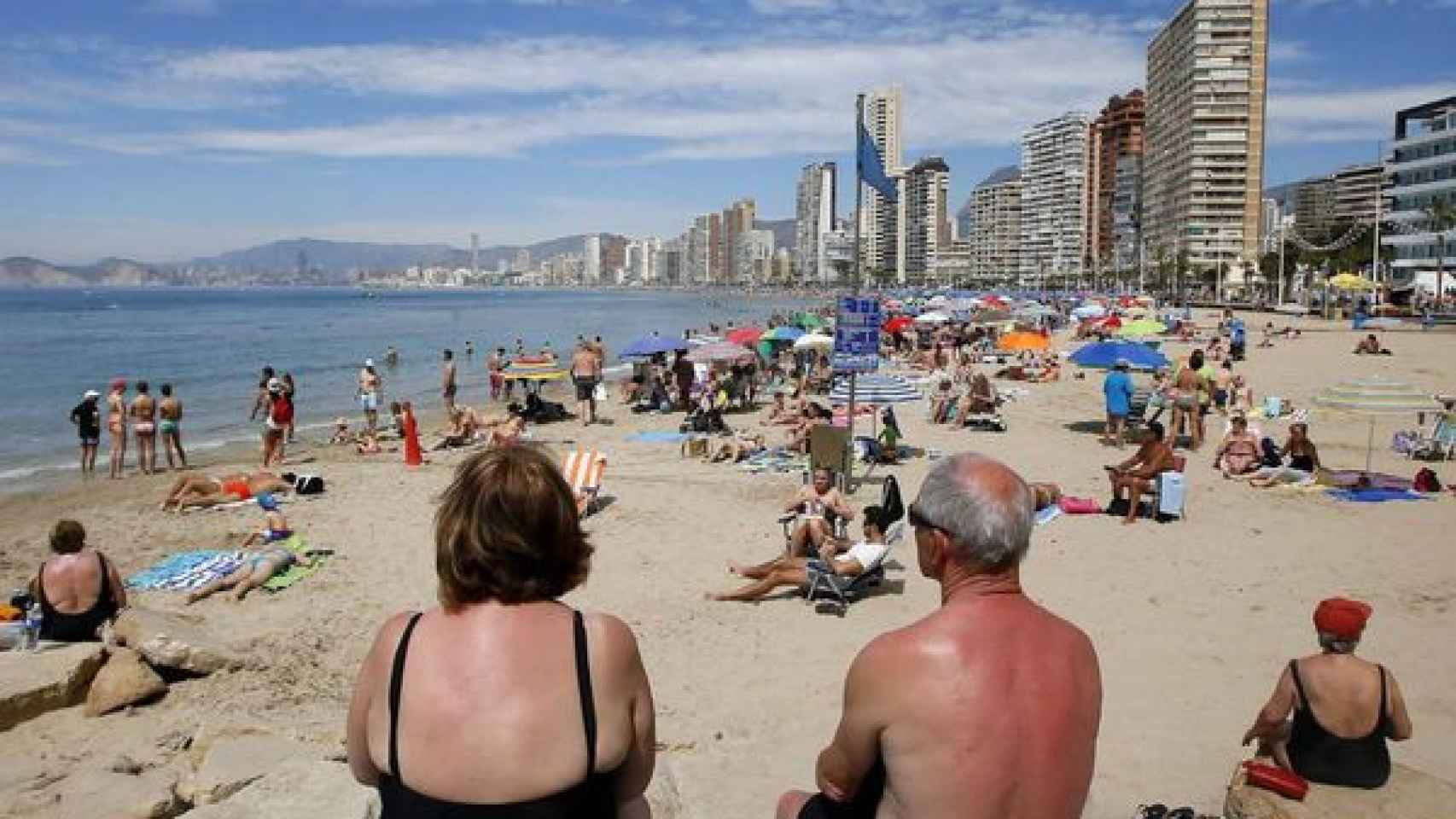 Dos personas mayores miran sentadas la línea de playa de Benidorm