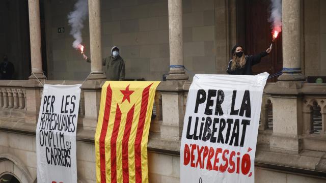 Dos personas sostienen bengalas junto a dos pancartas y una bandera estelada, durante una manifestación en el edificio histórico de la Universitat de Barcelona (UB)