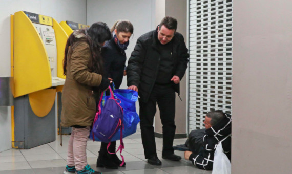 Dos voluntarias con el párroco repartiendo ropa a un indigente que conocen en un cajero / A.V.