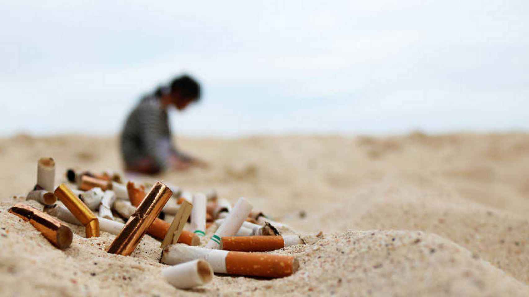 Decenas de colillas en la arena de la playa con una persona en el fondo