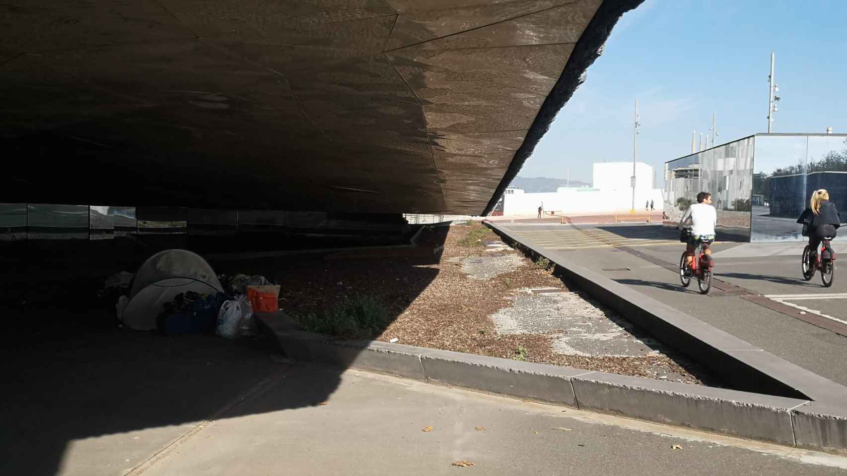 Dos ciclistas pasan por delante de una tienda donde duerme una persona en el Fòrum / GUILLEM ANDRÉS