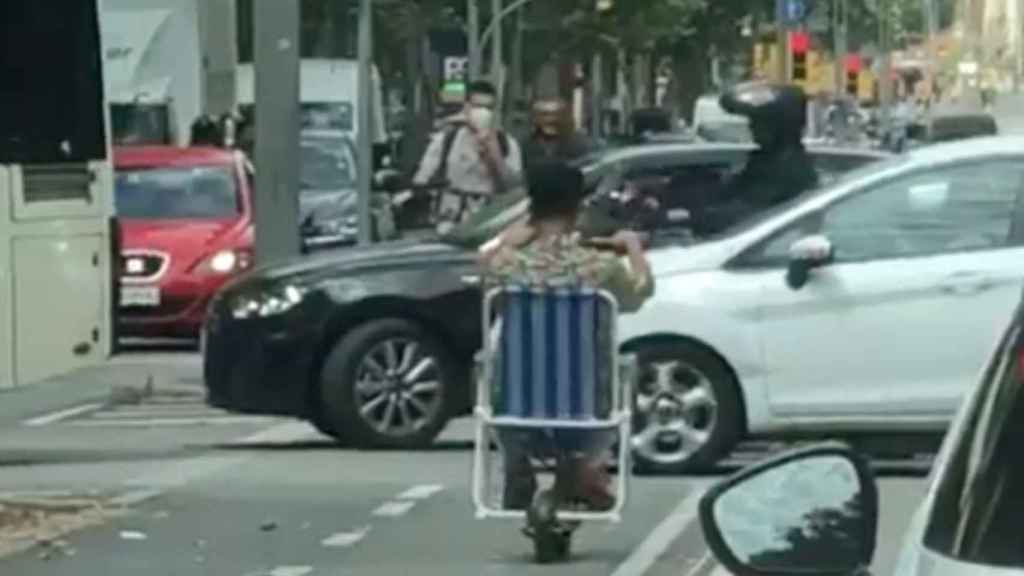 Un joven circula con un patinete eléctrica sentado encima de una silla de playa / @SosPobleSec