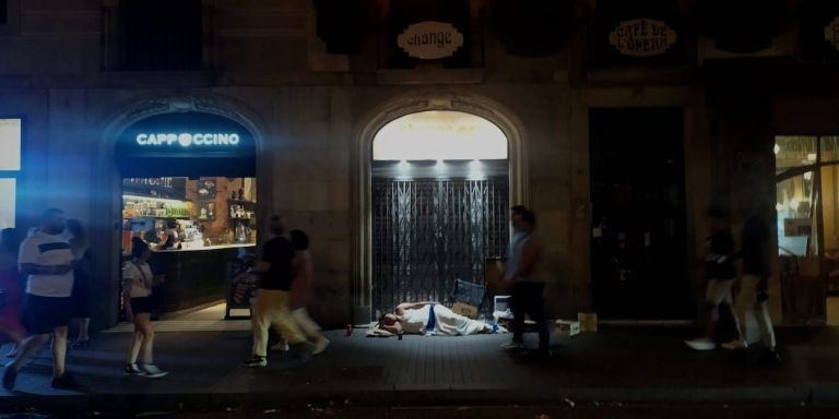 Una persona sintecho duerme en la Rambla mientras unos transeúntes pasan por su lado / PABLO MIRANZO - MA