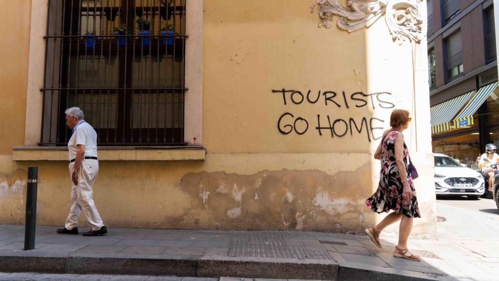 Vecinos paseando por una calle con un grafiti contra el turismo de fondo